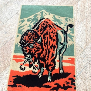 May include: Rectangular rug featuring a stylized bison design. The bison is depicted in shades of orange and black against a backdrop of a mountain and sky in beige and teal. The rug has a textured appearance.