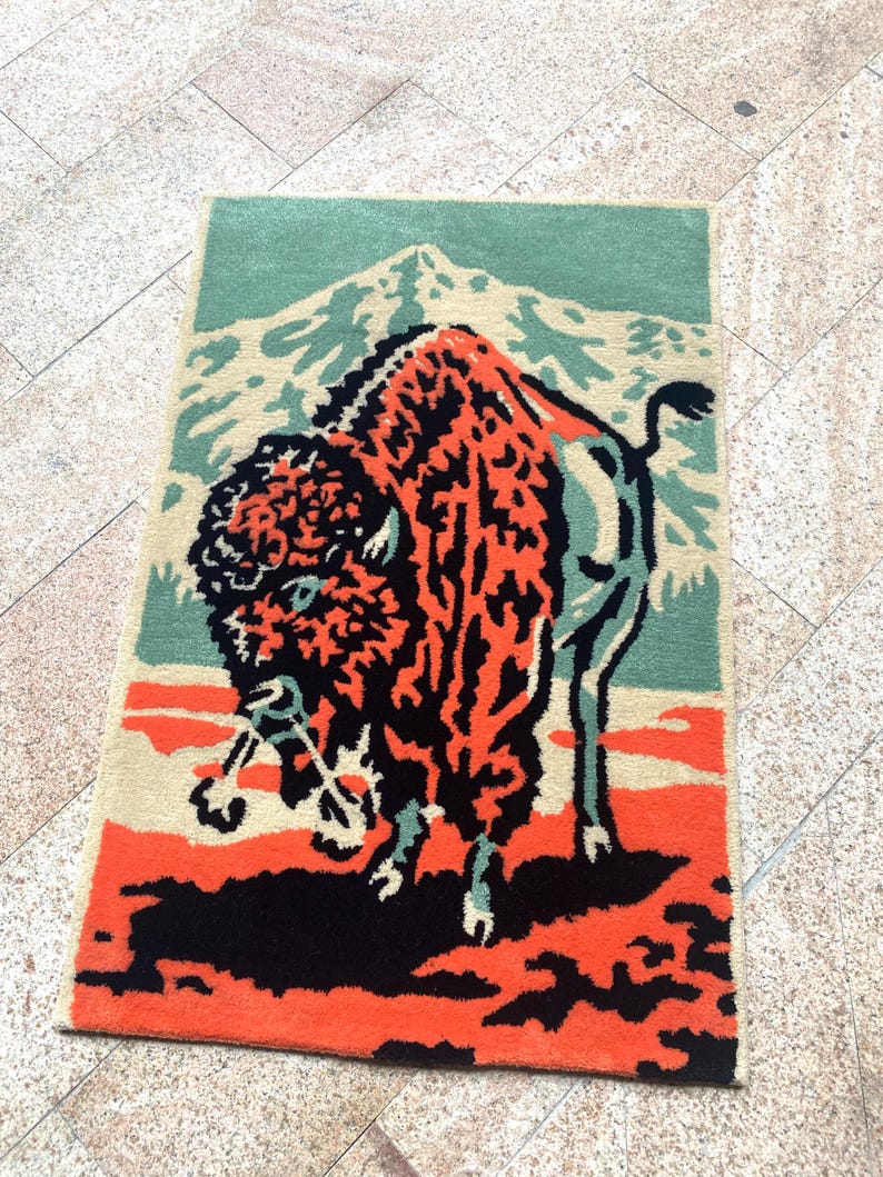 May include: Rectangular rug featuring a stylized bison design. The bison is depicted in shades of orange and black against a backdrop of a mountain and sky in beige and teal. The rug has a textured appearance.