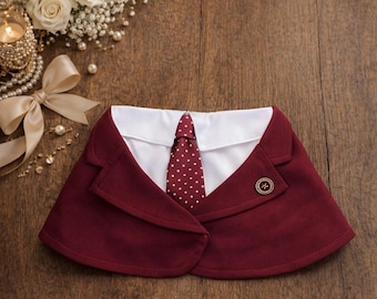 May include: A burgundy blazer collar with a white shirt collar underneath and a burgundy necktie featuring white polka dots. The blazer features a single round gold-toned button with a star emblem on the front lapel, creating a formal appearance.