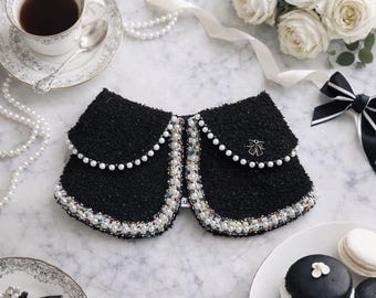May include: A black tweed collar with pearl and iridescent bead trim. The collar has a pearl chain detail and a small silver-tone floral embellishment. The image also includes a cup of tea, white roses, and macarons.