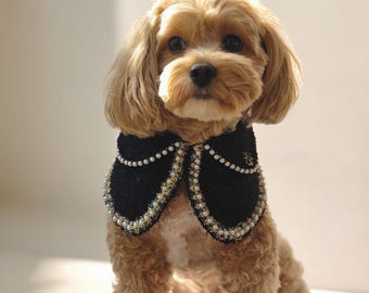 May include: A small, light brown dog wearing a black collar adorned with white pearls and iridescent beads. The collar has a decorative design and a small gold emblem. The text "LUKO LUKO" is visible at the bottom.