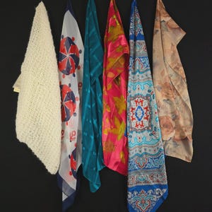 May include: A collection of six scarves hanging vertically. The first is a cream knit scarf, followed by a white scarf with red and blue circular patterns, a solid teal scarf, a pink scarf with gold leaf prints, a blue paisley scarf, and a beige floral scarf.