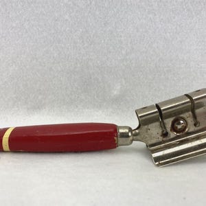 May include: A vintage cheese slicer with a red wooden handle and a stainless steel cutting head. The handle has a small hole at the end and a thin yellow stripe near the metal connection. The cutting head has multiple slots.