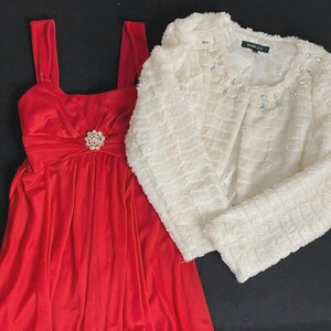 Puede incluir: Un vestido rojo con un lazo y una broche decorativa. Una chaqueta blanca texturizada con adornos de joyas está al lado del vestido. Las prendas se muestran sobre una superficie negra.