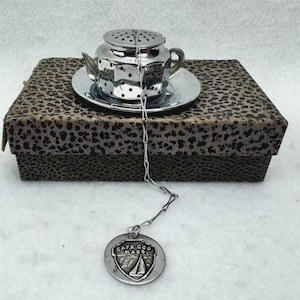 May include: A silver-toned metal tea infuser shaped like a miniature teapot on a matching saucer, connected by a chain to a round charm featuring a sailboat emblem and the text CAPE COD MASS. The set rests on a rectangular box with a leopard print pattern.