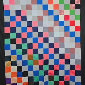 May include: A colorful patchwork quilt with a checkerboard pattern. The squares are in various colors, including pink, blue, green, gray, and red. The quilt is made of different fabrics and textures, creating a visually interesting design.