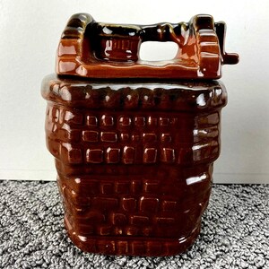 May include: A brown ceramic cookie jar with a lid and handle. The jar is shaped like a basket with a brick pattern. The lid has a handle and a small lever. The jar is a dark brown color.