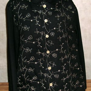 May include: Black button-down jacket with long sleeves. The jacket features an embroidered floral design in a light beige color. The buttons are round and beige. The jacket is displayed on a hanger.