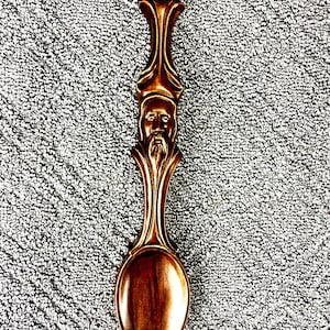 May include: A decorative, bronze-colored spoon with a detailed handle featuring a bearded face and ornate design. The spoon's bowl is oval-shaped. The spoon is likely a collectible or decorative item.