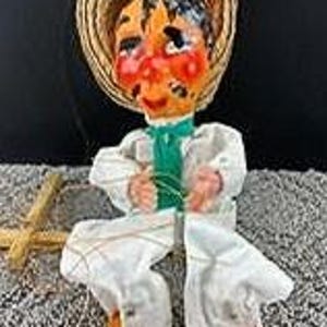 May include: A vintage puppet doll with a straw hat, white outfit, and orange shoes. The doll has a painted face with rosy cheeks and a green scarf. It is sitting with its hands clasped, and a wooden cross is visible.