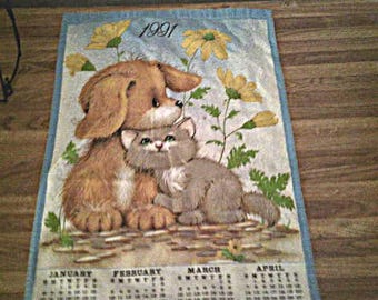 May include: A vintage calendar featuring a brown puppy and a gray kitten cuddling together. The calendar has a light blue border and includes the year 1991, yellow flowers, and the months January through April.