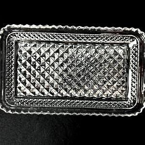 May include: A rectangular, clear glass serving tray with a diamond pattern in the center and a scalloped edge. The tray has two curved handles on either side. The tray is likely used for serving food or displaying items.
