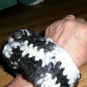 May include: A black, white, and gray crocheted wrist cuff with a decorative silver button. The cuff is wrapped around a wrist. The crochet pattern is a mix of black, white, and gray yarn, creating a textured design.