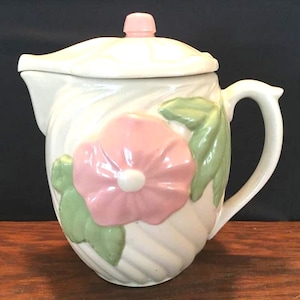 May include: Cream-colored ceramic teapot with a pink flower and green leaves design. The teapot has a matching lid with a pink knob and a curved handle. The body of the teapot has a textured, swirled pattern.
