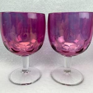 May include: A pair of iridescent magenta glass goblets featuring a rounded bowl with a textured pattern of circular indentations near the base. Each goblet has a clear glass stem and a circular foot, providing a stable base for the vessel.