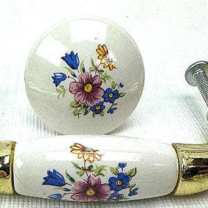 May include: A set of cabinet hardware featuring a gold-toned handle and a round knob. Both pieces have a white ceramic surface decorated with a floral pattern in pink, blue, and yellow.