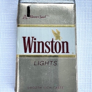 May include: A vintage Winston Lights cigarette pack in a gold and white color scheme. The pack features the Winston logo in burgundy, the word "LIGHTS", and the text "SMOOTH RICH TASTE". The top has two brown cylindrical pieces.