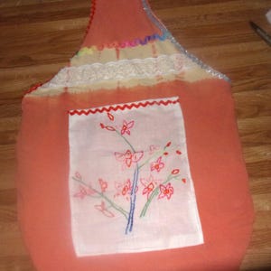 以下が含まれることがあります： ピンク色のトートバッグで、白いポケットにはピンクの花が刺繍されています。バッグにはレースのトリムと、上部にカラフルなトリムが付いています。