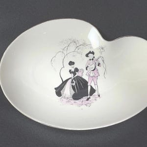 May include: White, kidney-shaped serving dish with a decorative illustration of two figures in period clothing. The figures are in black, pink, and gray. The dish has a silver rim and is set against a dark background.