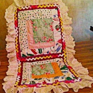 May include: A vintage patchwork quilt with a central square of floral and patterned fabrics, framed by a red and yellow zigzag border. The edges are finished with cream-colored lace trim. The quilt is handmade and has a traditional aesthetic.