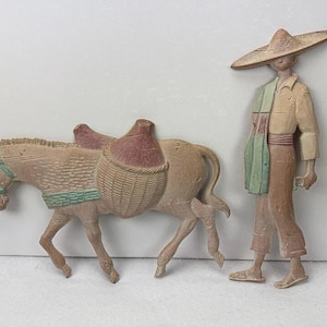 May include: A wall-mounted decorative set featuring a tan donkey with a green harness and a woven basket on its back, alongside a standing figure wearing a wide-brimmed hat, a collared shirt, trousers, and sandals, both rendered in muted earth tones.
