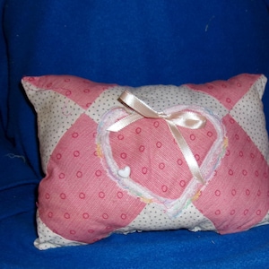 May include: A small, rectangular pillow with a patchwork design. The pillow features a pink heart in the center, adorned with a white ribbon bow and a small white heart-shaped button. The pillow's fabric is a mix of pink polka dots and white polka dots.