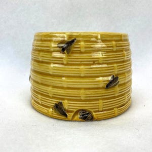 May include: A ceramic beehive-shaped container in a light yellow color, with a textured, woven design. The container has several small, dark-colored bee figurines attached to the exterior. The beehive has an opening at the bottom.
