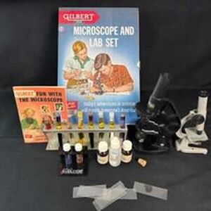 May include: A Gilbert Microscope and Lab Set featuring a large box with illustrations, a Fun with the Microscope manual, a black microscope, a white microscope, a test tube rack with glass vials, several small bottles, and clear glass slides.
