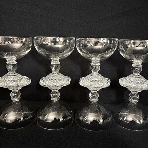 May include: Four clear, vintage champagne coupe glasses. Each glass features a wide, shallow bowl atop a decorative, textured stem and a rounded base. The glasses are arranged in a row, reflecting light.