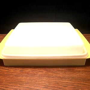 May include: A vintage, square-shaped food storage container with a white lid and a pale yellow base. The container has a rectangular shape with rounded corners. The lid sits flush with the base, creating a clean, classic look. The container is made of plastic.