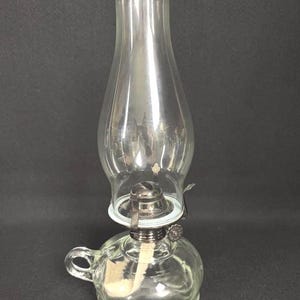 May include: A clear glass oil lamp featuring a rounded base with a small side handle, a silver-toned metal burner assembly with an adjustment knob, and a tall, bulbous clear glass chimney that sits securely atop the burner mechanism.