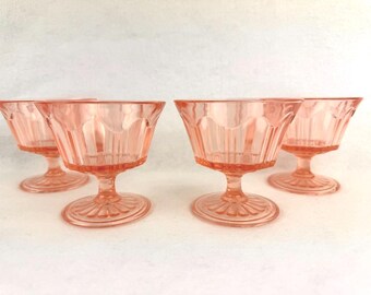 May include: Four vintage pink glass dessert or champagne glasses. Each glass has a stemmed base with a decorative design and a bowl with vertical and arched detailing. The glasses are arranged in a row against a white background.