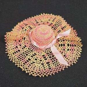 May include: A miniature doll hat crafted from crochet thread in a variegated pattern of soft pink and pale yellow. The hat features a rounded crown and a wide, scalloped brim with an openwork design, accented by a light pink satin ribbon tied.