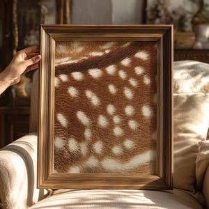 Puede incluir: Una obra de arte enmarcada que presenta un primer plano de piel de ciervo con un patrón de manchas marrones y blancas. El marco es de color marrón claro con un diseño acanalado. La obra de arte se exhibe en un sofá beige.