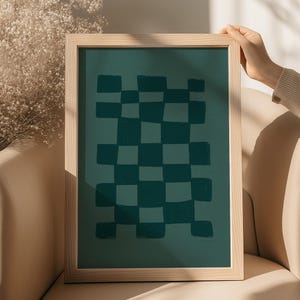 May include: A framed artwork featuring a teal checkerboard pattern. The artwork is in a light-colored wooden frame. The checkerboard design is composed of dark teal squares on a lighter teal background. The artwork is held by a person.