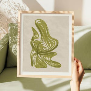May include: A framed wall art print featuring an abstract design of overlapping olive green lines on a light beige background. The artwork is enclosed in a light-colored wooden frame, showcasing a fluid, organic shape created by continuous strokes.