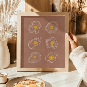 Peut inclure: Une affiche encadrée avec six illustrations d'œufs abstraits, avec des contours blancs et des jaunes d'œufs jaunes sur fond marron. Le cadre est en bois clair.