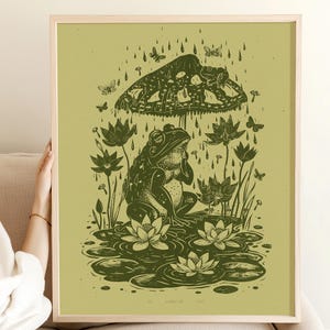 Op de afbeelding: Een ingelijste kunstprint met een illustratie van een kikker onder een paddenstoel, omringd door regen en vlinders. De illustratie, in groentinten, bevat waterlelies en bloemen, wat een grillige, vintage-achtige scène creëert.