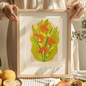 Op de afbeelding: Een ingelijste kunstdruk met een botanische illustratie van garnalen, citroenschijfjes en groene bladeren. Het kunstwerk heeft een lichtbeige achtergrond en is ingelijst in een licht houten frame. De tekst onderaan luidt "Aromatic & succulent BOTANICAL SHRIMPS."