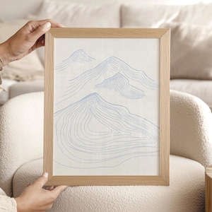 May include: A framed art print featuring a minimalist mountain design. The artwork showcases blue contour lines on a cream-colored background, encased in a light brown wooden frame. The piece is held by a person.