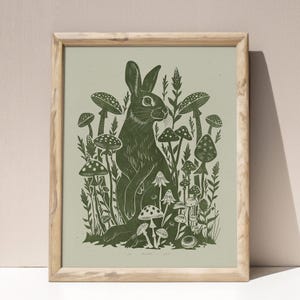 Puede incluir: Impresión enmarcada con una ilustración detallada en linóleo de un conejo rodeado de setas y follaje. La obra de arte está realizada en tonos verdes sobre un fondo verde claro, con un marco de madera natural.