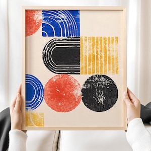 Op de afbeelding: Abstracte kunstafdruk in een licht houten frame. Het kunstwerk toont geometrische vormen in rood, blauw, zwart en geel op een crèmekleurige achtergrond. Het ontwerp omvat cirkels, bogen en rechthoekige vormen.