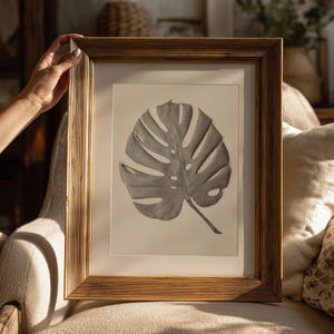 May include: A framed botanical print featuring a gray monstera leaf. The leaf is centered on a cream-colored background, encased in a gold-toned, rectangular frame. The frame has a textured, ridged design. The print is held by a person.