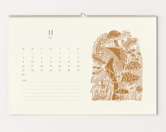 May include: A cream-colored calendar with a spiral binding, featuring the month of November. The right side showcases a detailed illustration of various mushrooms and foliage in shades of brown. The calendar includes the days of the week and dates.