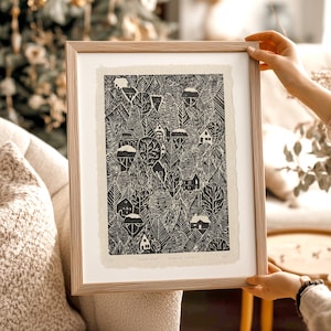 May include: A framed black and white art print depicting a forest scene with houses nestled among the trees. The artwork is in a light-colored wooden frame. The print has a textured, handmade paper appearance.