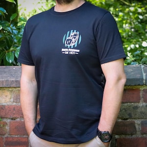 Może przedstawiać: Czarny t-shirt z okrągłym dekoltem z grafiką na piersi. Grafika przedstawia stylizowany wzór kasku do futbolu wewnątrz niebiesko-białego koła w paski, z napisem "Marine Maintenance - Est 1871".