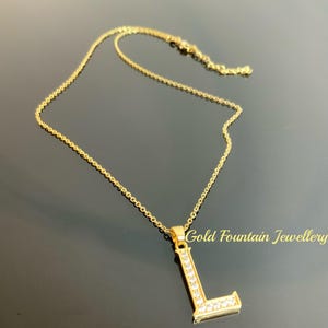 May include: A gold chain necklace with a pendant in the shape of the letter L. The letter is encrusted with small clear stones. The necklace is on a black background. Gold Fountain Jewellery is written in white text on the bottom right corner of the image.