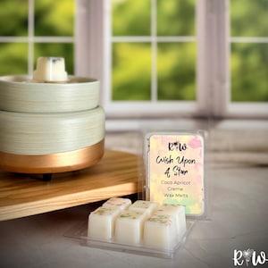 May include: A two-tiered, cream-colored wax warmer with a copper-colored band, and a wooden base. A package of Coco Apricot Creme wax melts is next to the warmer. The text on the package reads "Wish Upon A Star".