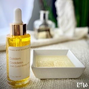 May include: A clear glass bottle of Golden Goddess 24K Gold Body Oil, with a gold-colored dropper and cap. The bottle is next to a small white square dish filled with the same golden oil. The label reads "Roses and Wildflowers".