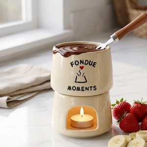 May include: A cream-colored ceramic fondue pot with a brown wooden-handled fork. The pot features the text FONDUE MOMENTS and a small red heart icon. It is filled with melted chocolate and sits on a base containing a lit white tea light candle.